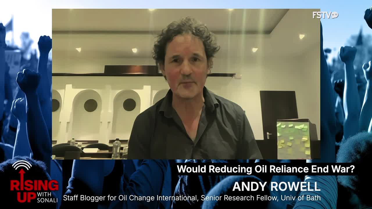 A man with curly hair is speaking on Free Speech TV. Behind him, a crowd of people with raised fists fills the screen.