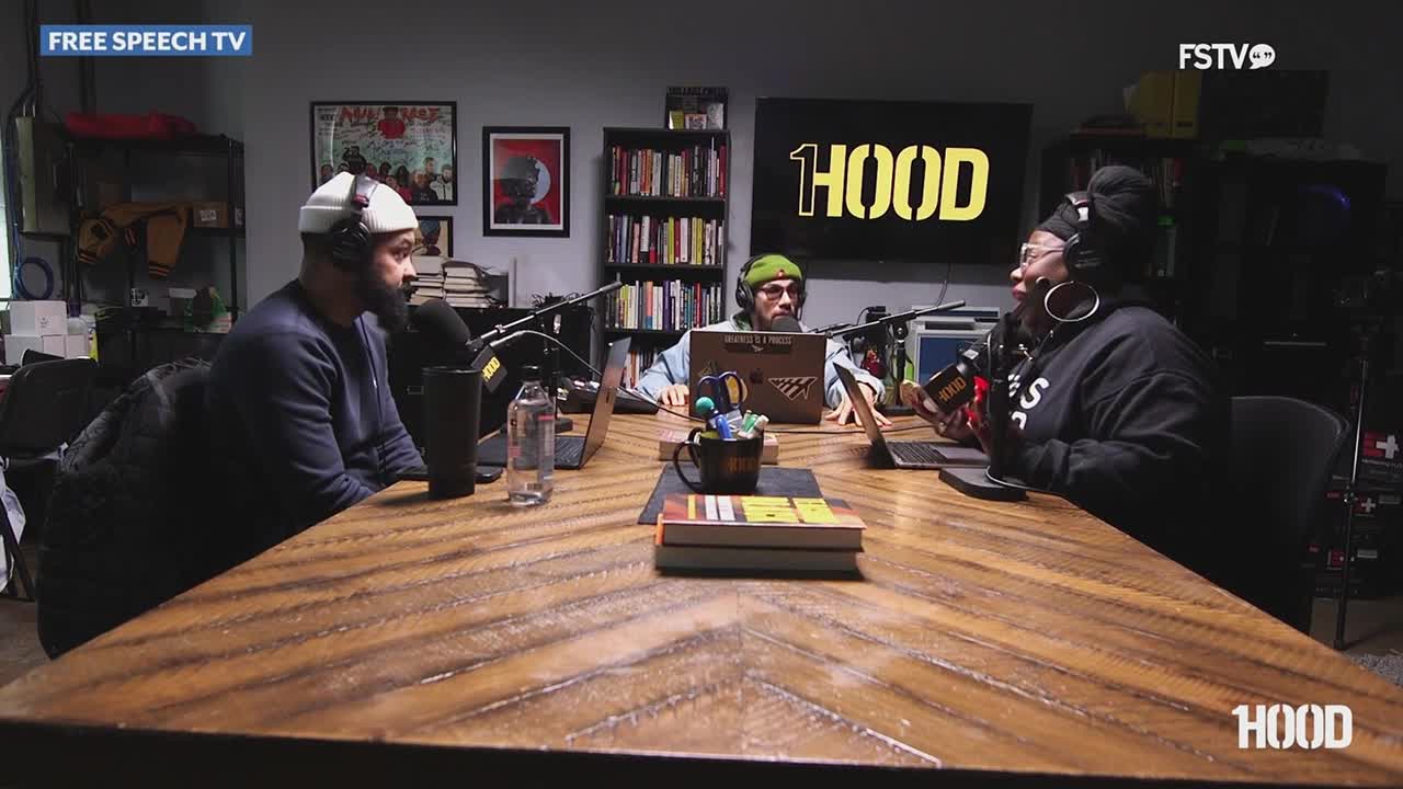Three people sit around a wooden table in a studio, microphones in front of each of them. The "1HOOD" logo is displayed prominently on a screen behind them. This is a recording session for Free Speech TV.