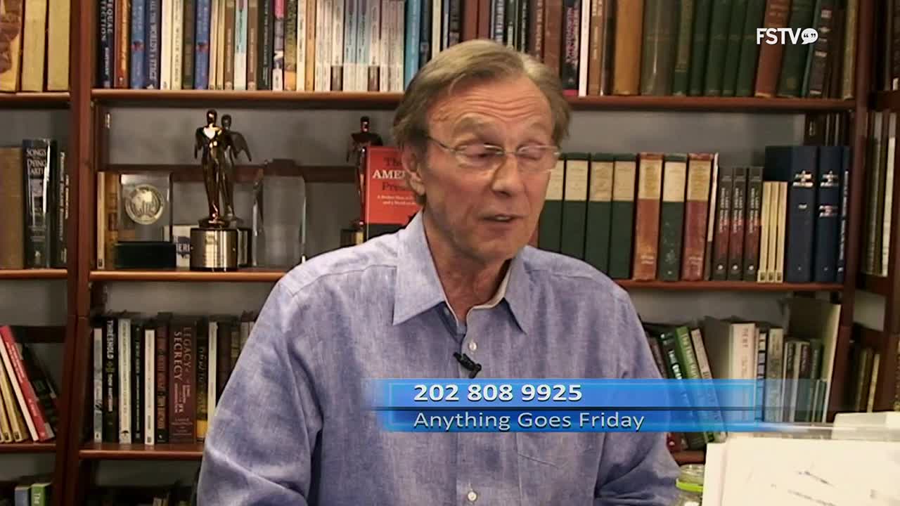 A man in a blue button-down shirt speaks, his glasses reflecting the light. Behind him, shelves packed with books stretch upwards, a testament to countless hours of reading and research.