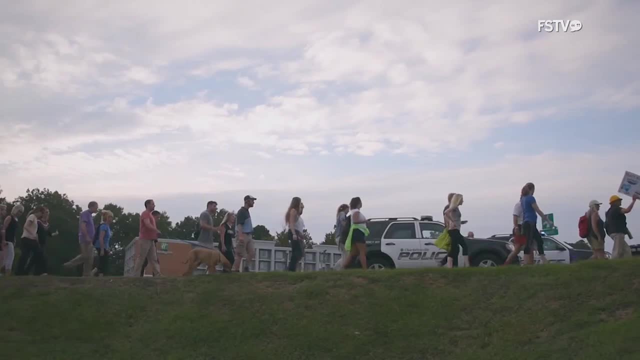 A group of people walks along a grassy embankment, some holding signs. A police car follows them, its lights off.