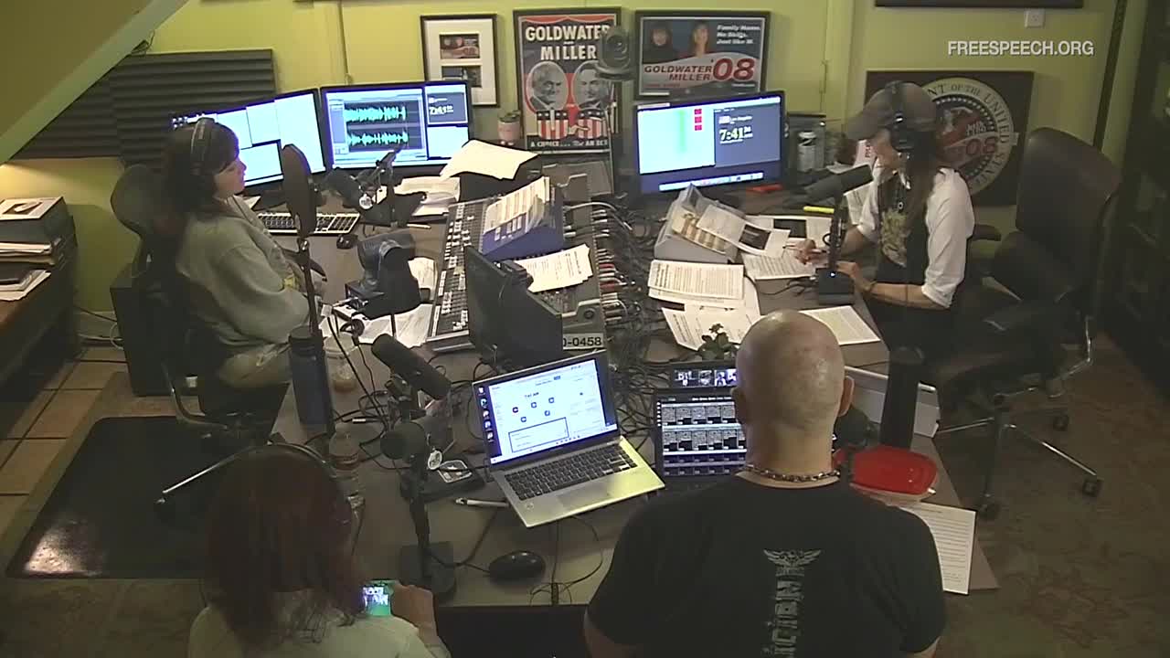 A woman in headphones speaks into a microphone at a cluttered desk, while another person, seen from behind, faces a bank of monitors. Papers and audio equipment fill the space, with a "freespeech.org" logo visible on the wall.