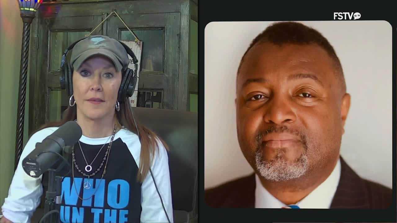 A woman with headphones and a baseball cap speaks into a microphone, her t-shirt displaying "WHO RUN THE..." in large blue letters. To her right, a man in a suit and tie looks directly forward.