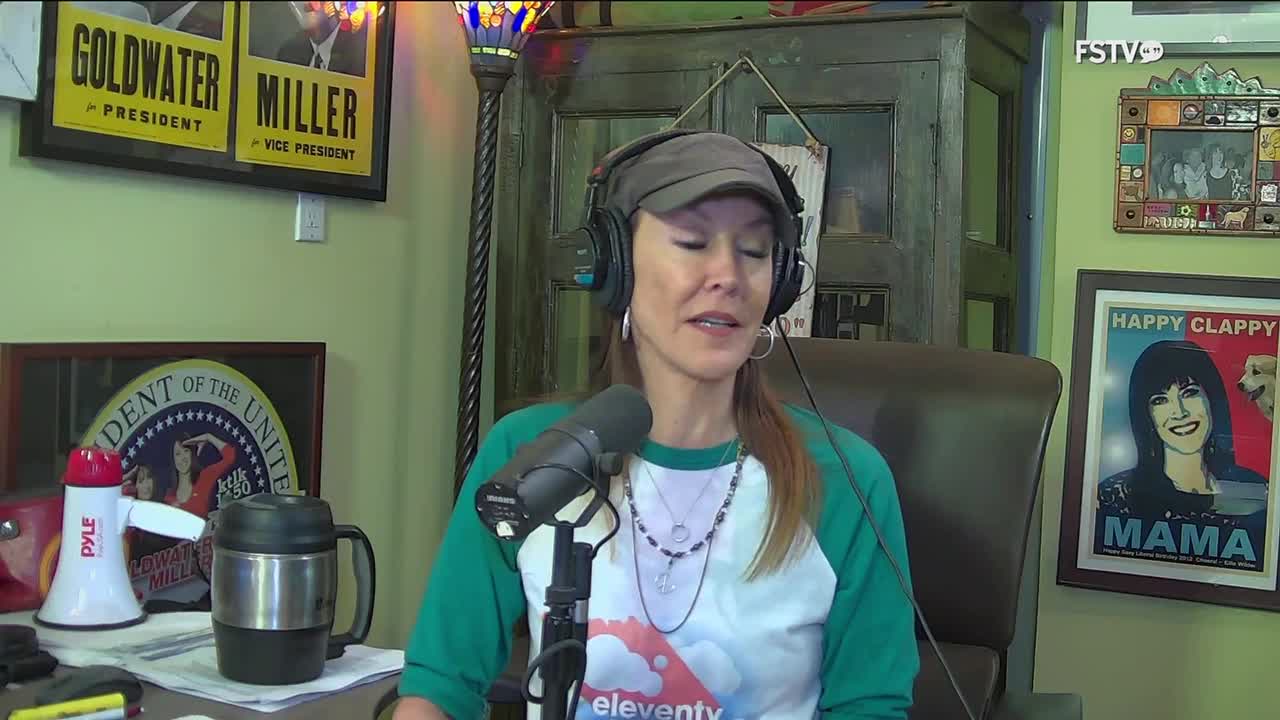 A woman with headphones on speaks into a microphone, her voice likely broadcast on Free Speech TV. Behind her, vintage political posters and a framed portrait of a woman with a dog adorn the wall.