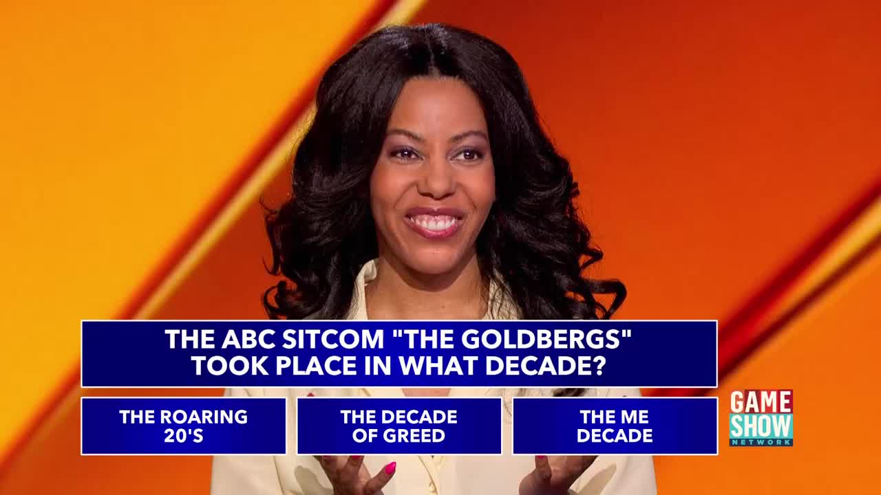 A smiling woman stands before a question on a Game Show Network set. The screen displays a question about the ABC sitcom "The Goldbergs," with three possible answers below.
