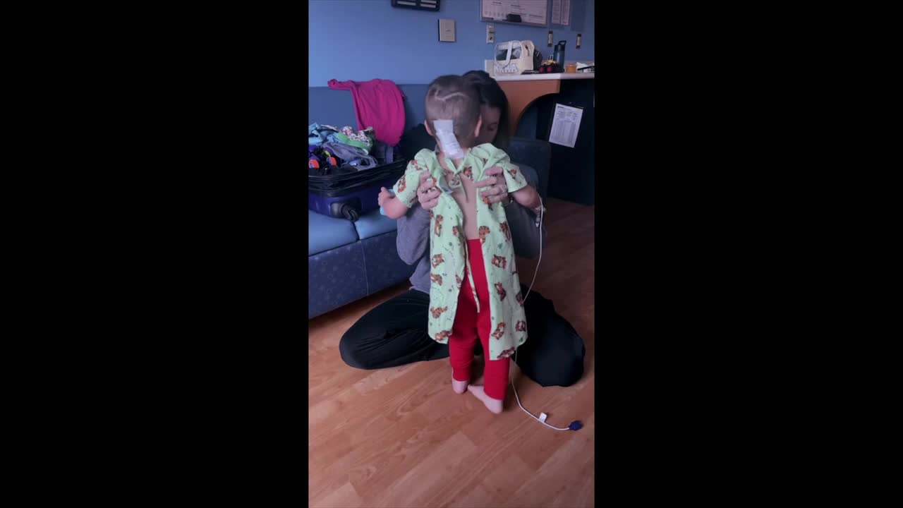 A small child, wearing red pants and a patterned robe, is being supported by an adult. A tube runs from the child's nose, and they're standing on their toes.
