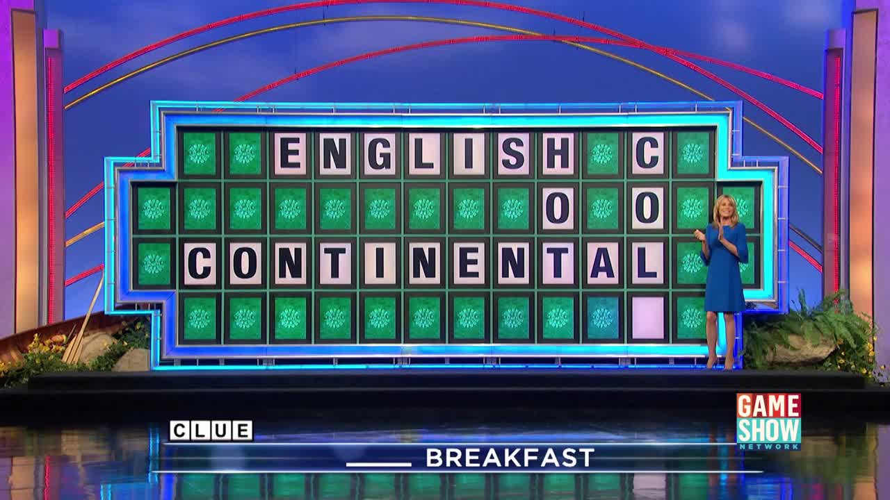 On the Game Show Network set, a woman in a blue dress stands beside a large puzzle board, revealing the letters of "Continental Breakfast." The clue below the board reads "BREAKFAST."
