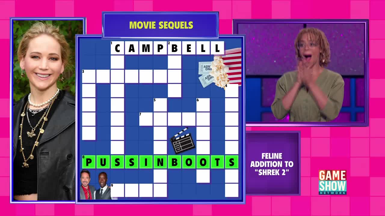 A woman on the Game Show Network beams with excitement, her hands clasped in front of her. The crossword puzzle board displays the answer "Puss in Boots" with a clue about a "Feline Addition to 'Shrek 2.'"
