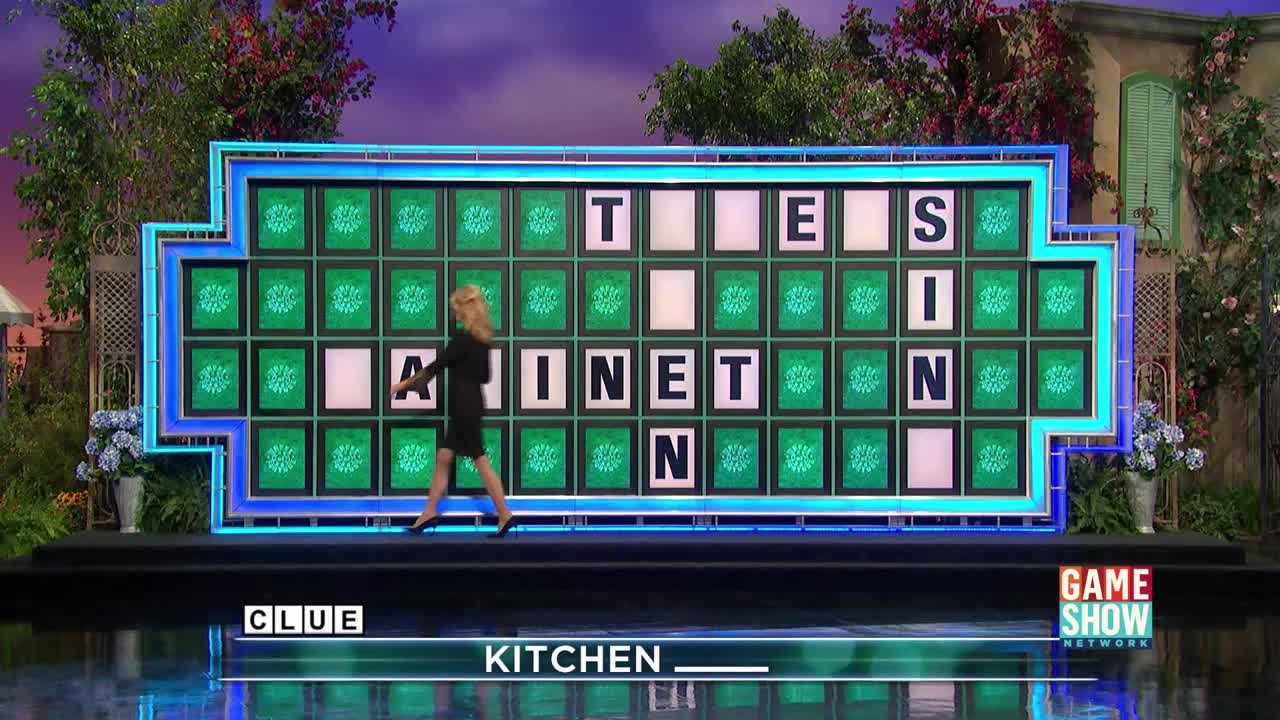 A woman in a black dress strides across the stage, the partially revealed puzzle board of a popular television game show behind her. The "Game Show Network" logo is visible in the lower right corner.
