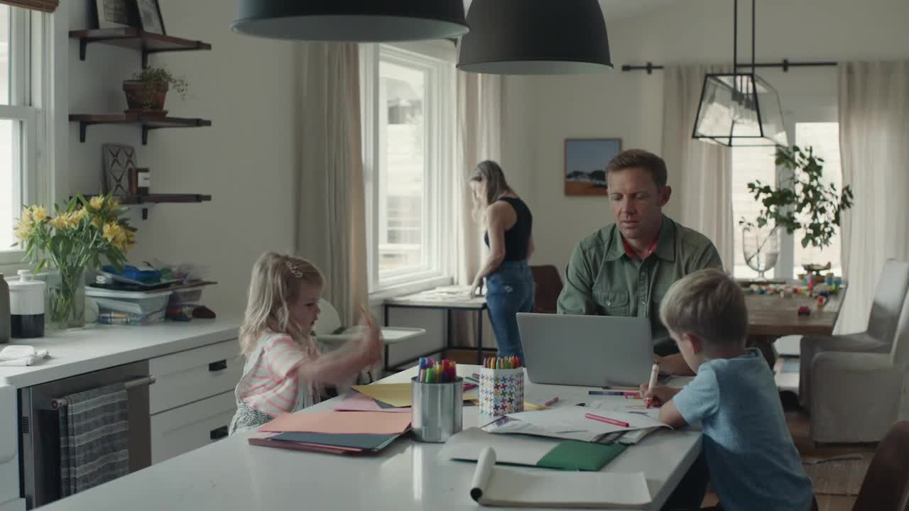 A man works on a laptop while his son colors at the kitchen island. A young girl waves her hands in the air, and a woman stands near the window.
