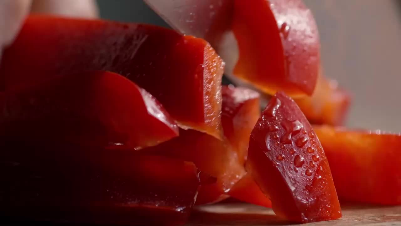 A chef's knife slices through a vibrant red pepper, breaking it into chunks. The glistening, freshly cut pieces are scattered on a wooden surface.
