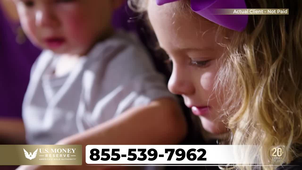 Two young girls are focused on something just out of view, one with a purple bow in her hair. A phone number for US Money Reserve is prominently displayed at the bottom of the screen.
