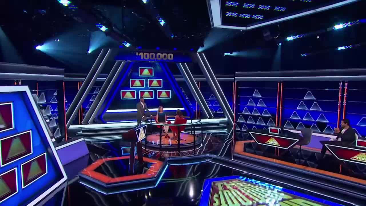 On the set of a game show, a host stands at a podium while two contestants sit at a circular table. The stage is lit with blue and white lights, and the backdrop features a pyramid of triangles.

