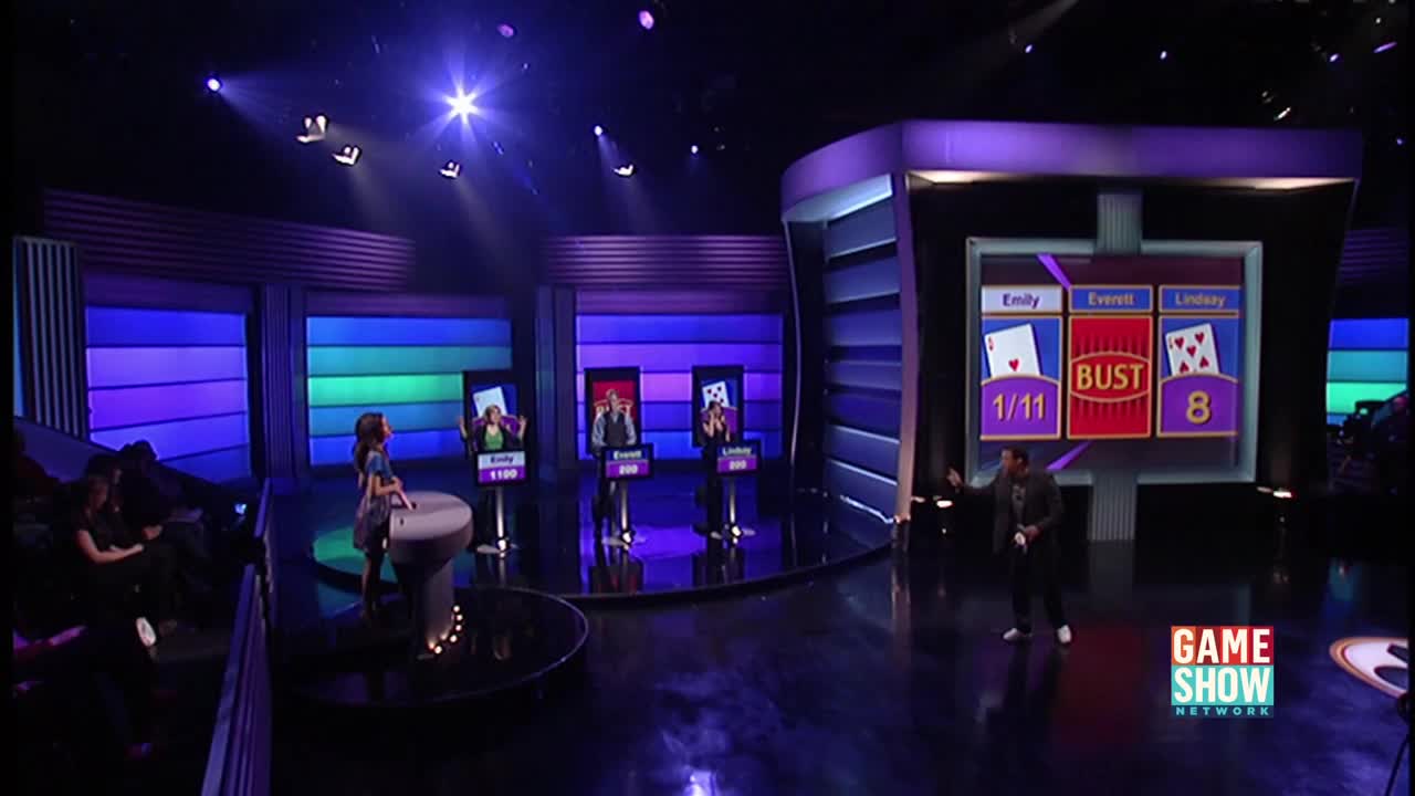 On a Game Show Network set, a host gestures toward a screen displaying card hands and the word "BUST." Three contestants stand at podiums, each lit by spotlights.
