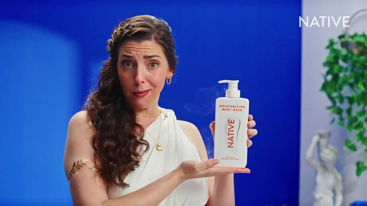 A woman in a toga-like outfit holds up a bottle of body wash, looking directly at the camera with a slightly surprised expression. Behind her, a bright blue backdrop contrasts with the white bottle and the greenery in the background.
