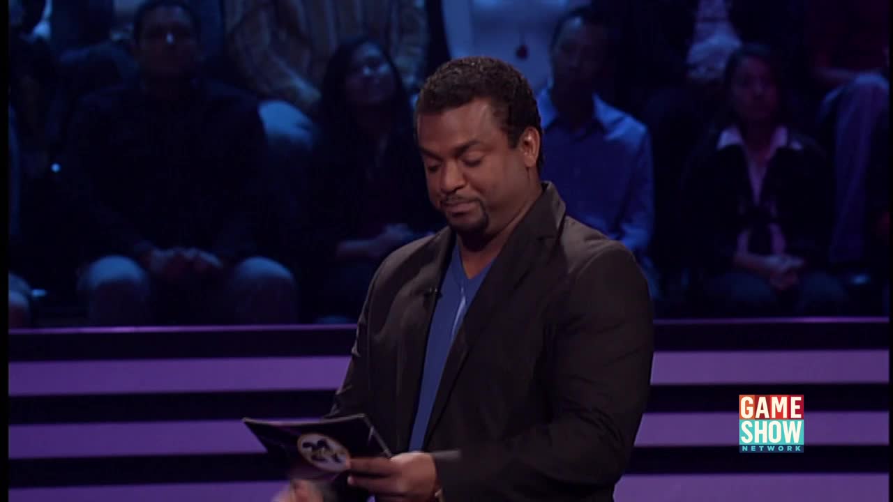 The host, on the Game Show Network, is reading from a card, his expression serious. A studio audience sits in the background, watching the unfolding scene.
