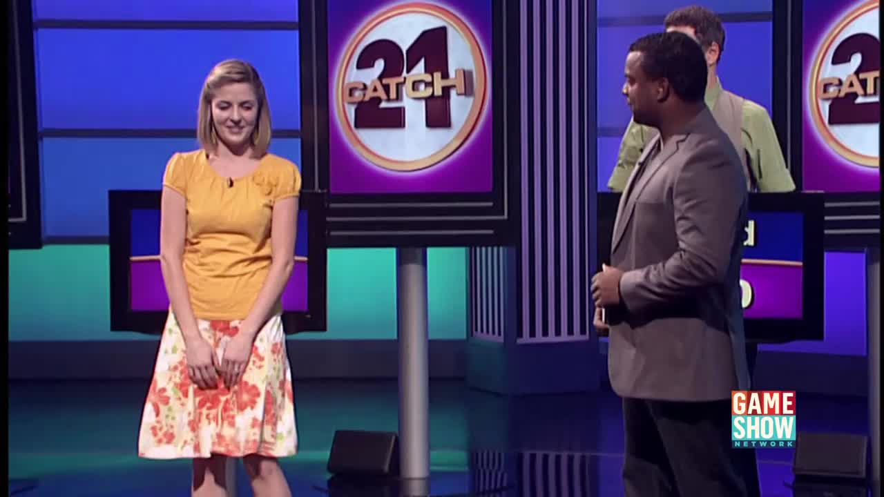 A woman in a floral skirt and yellow top stands on the set of *Catch 21*, looking down. The host, in a grey suit, is turned towards her, while another contestant stands behind him.
