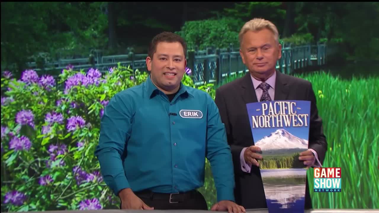 Erik, wearing a teal shirt and a name tag, smiles at the camera. A man in a suit holds a "Pacific Northwest" travel brochure, the Game Show Network logo visible in the corner.

