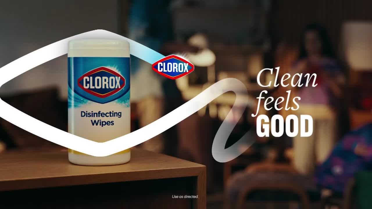 A Clorox Disinfecting Wipes container sits on a wooden surface, the brand name and product description clearly visible. A stylized white swirl and the words "Clean feels GOOD" float in the background, suggesting a comfortable domestic scene.
