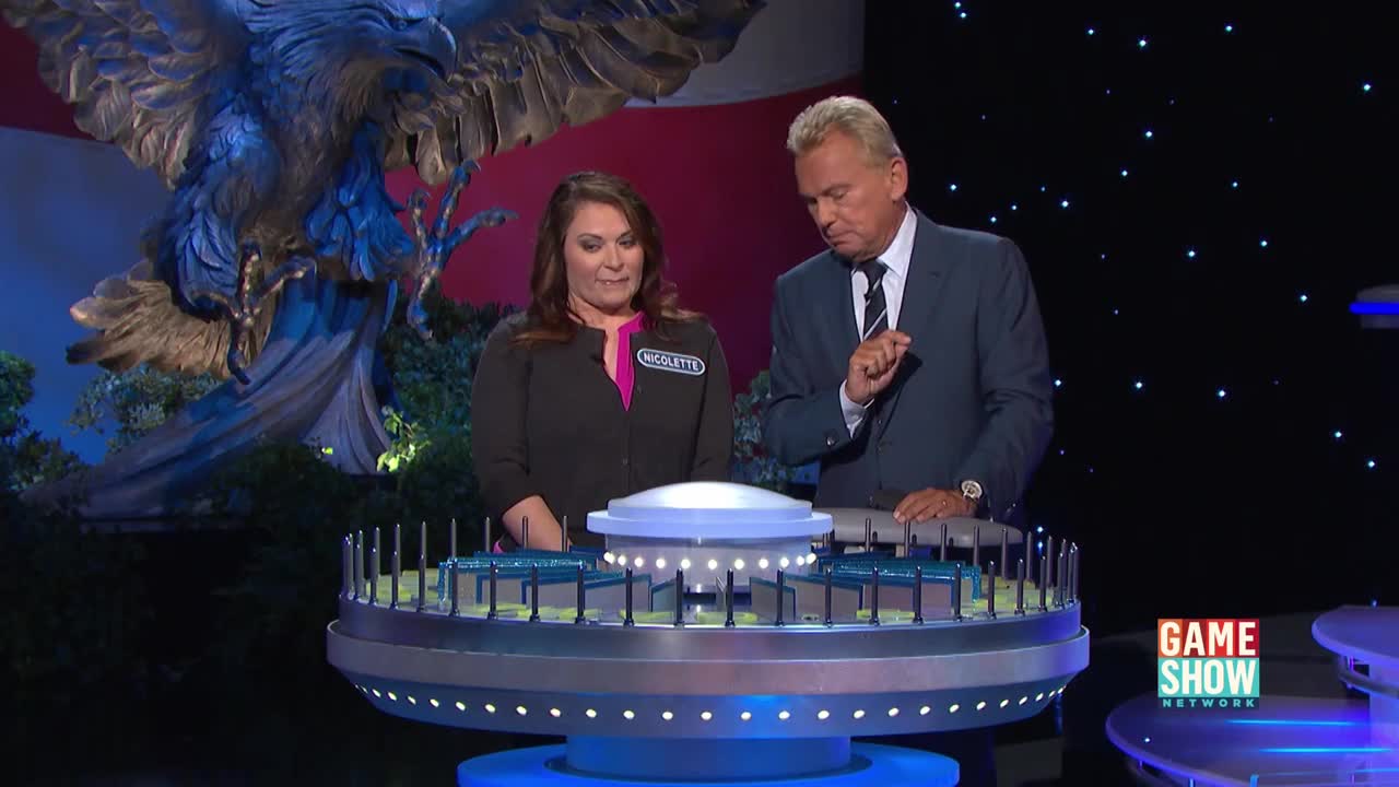 The woman, "Nicolette," looks intently at the game board as the host gestures towards it. A large, illuminated eagle statue stands behind them on the set of what appears to be a game show.
