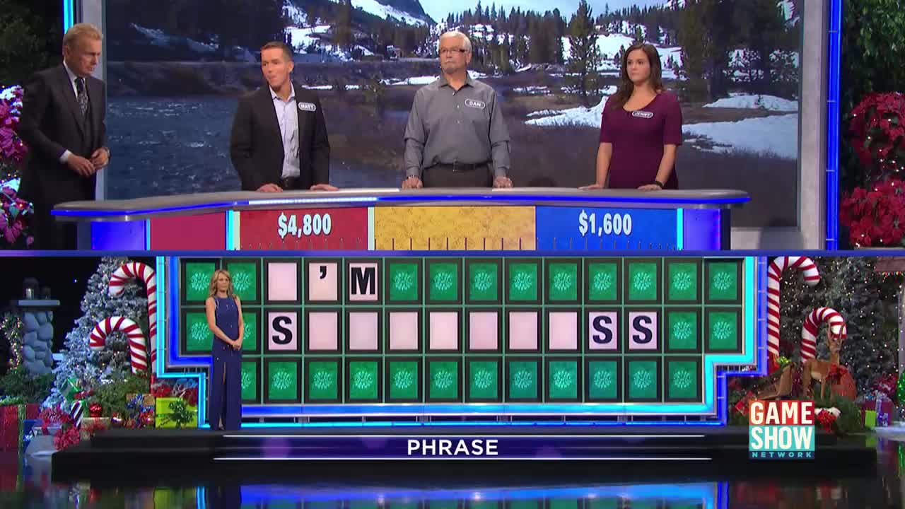 The host is standing at the wheel, and the contestants are waiting behind the podiums.  The board shows the letters of the phrase, and the final "S" is revealed.
