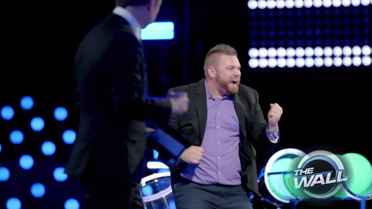 The man in the purple shirt throws his fists in the air, yelling with excitement. The other man in the suit seems to be reacting to the same moment.
