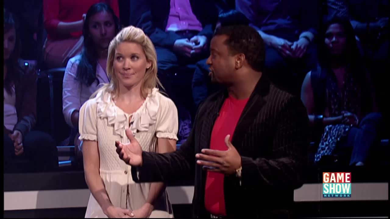 The host gestures with his hands as he speaks, facing the contestant. She stands beside him, smiling slightly, as the audience watches from the background.
