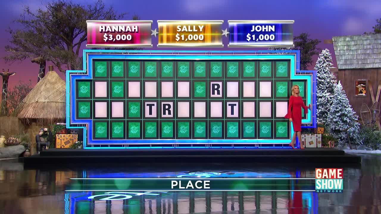 The host, in a red dress, stands near the "Wheel," which displays the letters "T," "R," and "T." The game show's backdrop is a mix of a safari and a snowy cabin.
