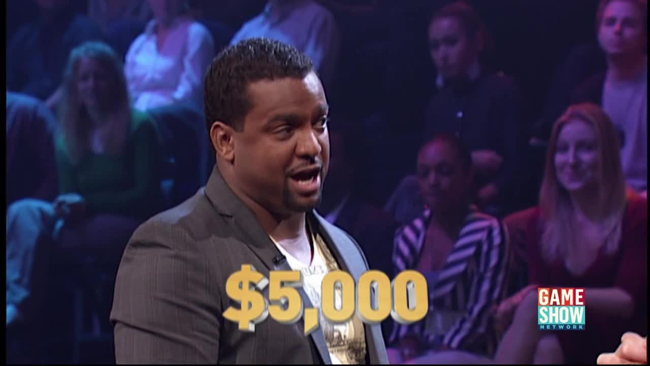 The man in the gray jacket is speaking, his mouth open. A graphic displaying "$5,000" floats in front of him, and the Game Show Network logo is visible.
