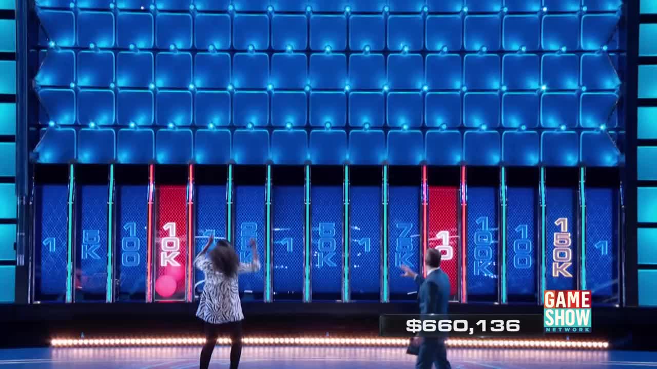 A woman in a zebra-print jacket and a man in a suit gesture at the large, glowing wall. Pink balls are dropping in front of the "10K" slot on the Game Show Network set.
