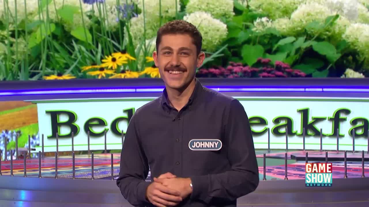 Johnny, wearing a name tag, smiles at the camera on a set decorated with flowers and the words "Bed... Breakfast." The Game Show Network logo is visible in the lower right corner.
