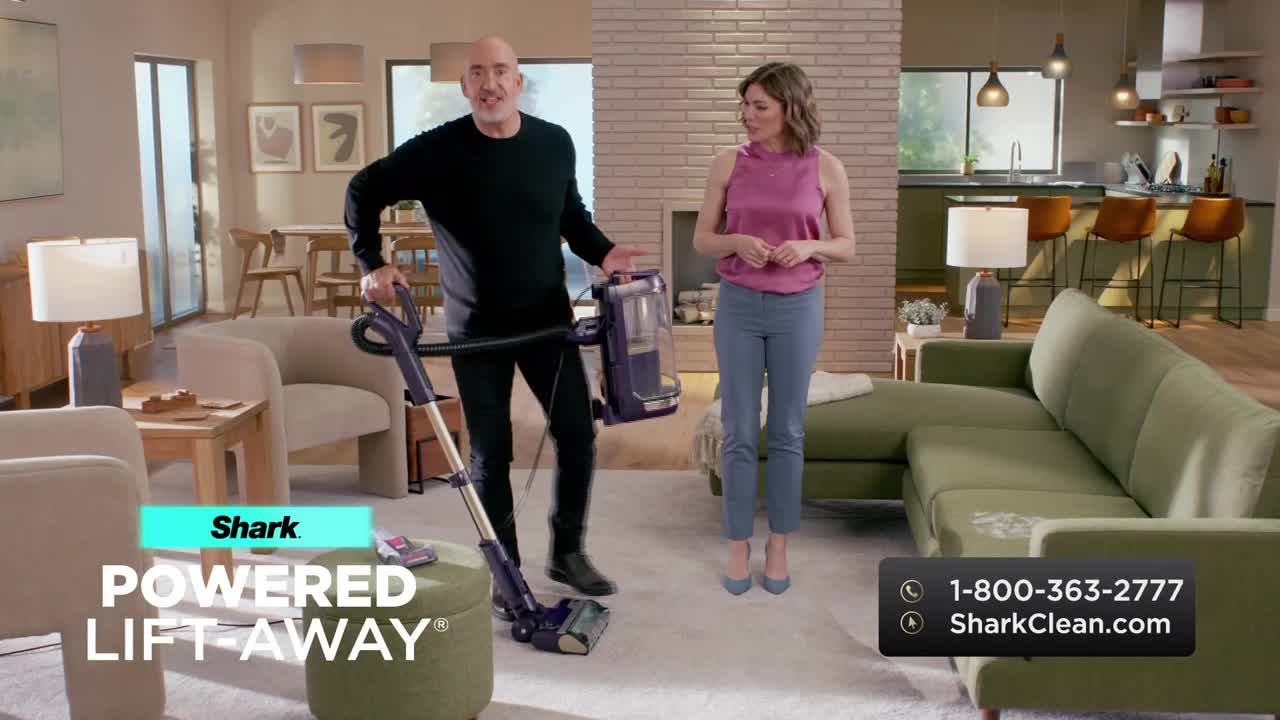 A man in black is holding a vacuum cleaner, gesturing towards the machine while a woman in a pink top stands nearby. The scene is a brightly lit living room, likely a set for a commercial airing on the Game Show Network.
