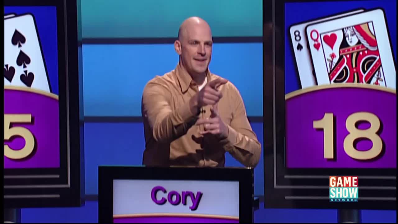 Cory, on a Game Show Network set, is pointing with his right hand. He's making a gesture with his left hand, and the cards and numbers are displayed behind him.
