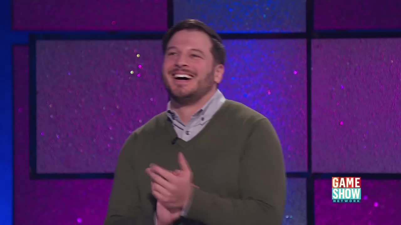 He's clapping, beaming, and laughing, clearly enjoying himself on the Game Show Network set. The background is a bright, geometric pattern, and the logo is visible in the corner.
