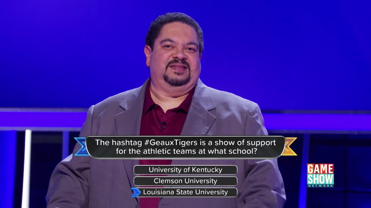 A man in a gray blazer stands in front of a blue backdrop, smiling as a question appears on the screen. The Game Show Network logo is visible in the corner, and the answer choices are listed beneath the question.
