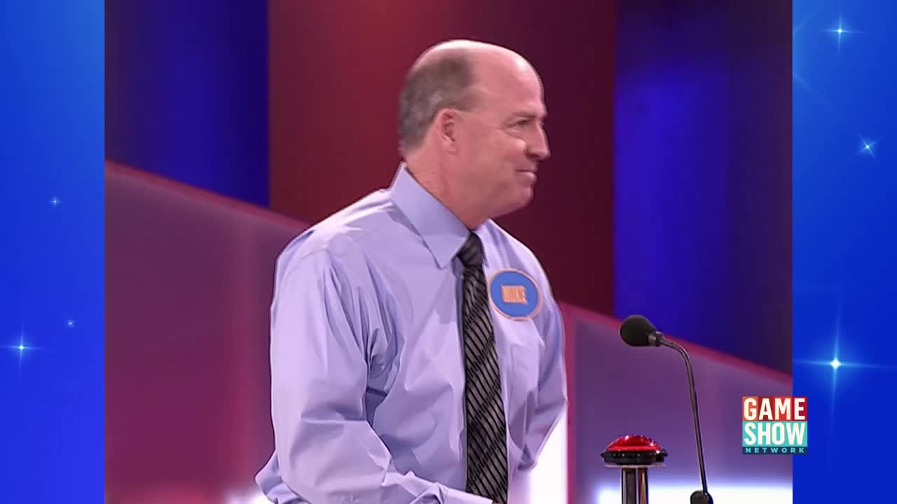Mike, wearing a name tag, stands at a podium on the Game Show Network set. He's looking to his right, likely waiting for the next question.
