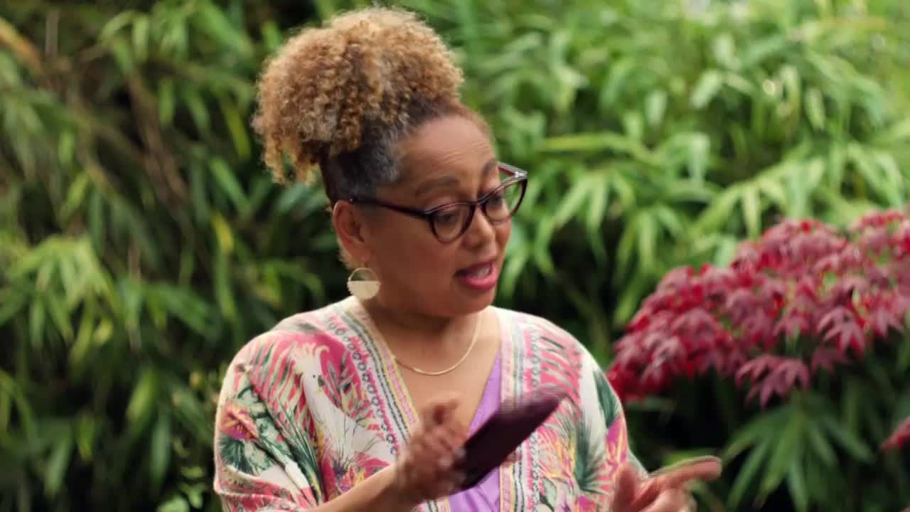 The woman in glasses gestures with her hands, holding a dark object. She's speaking, likely hosting a segment on the Game Show Network.

