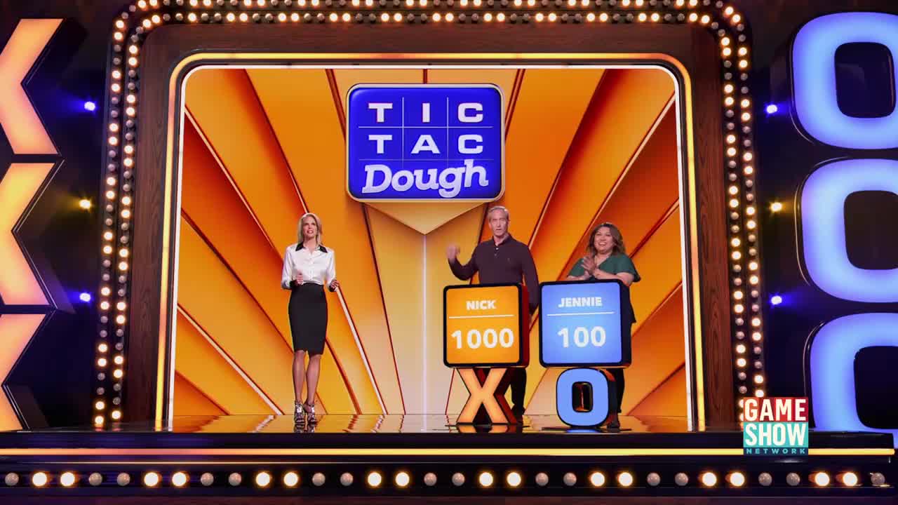 On the set of "Tic Tac Dough" on Game Show Network, the host gestures toward the contestants, who stand beside their scoreboards. The woman on the left stands still, while the other woman claps.
