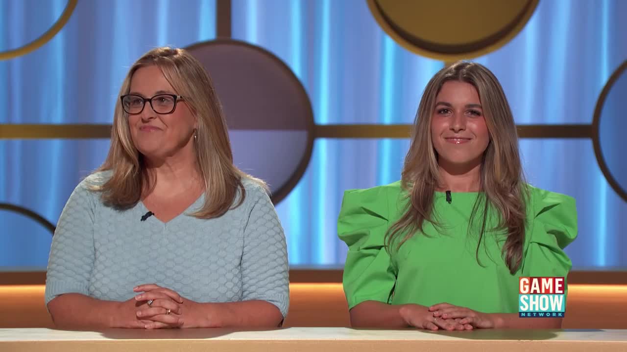On a Game Show Network set, two women sit side-by-side, each with their hands clasped in front of them. The woman on the right wears a bright green top, while the other is in a light blue sweater, both facing forward with pleasant expressions.
