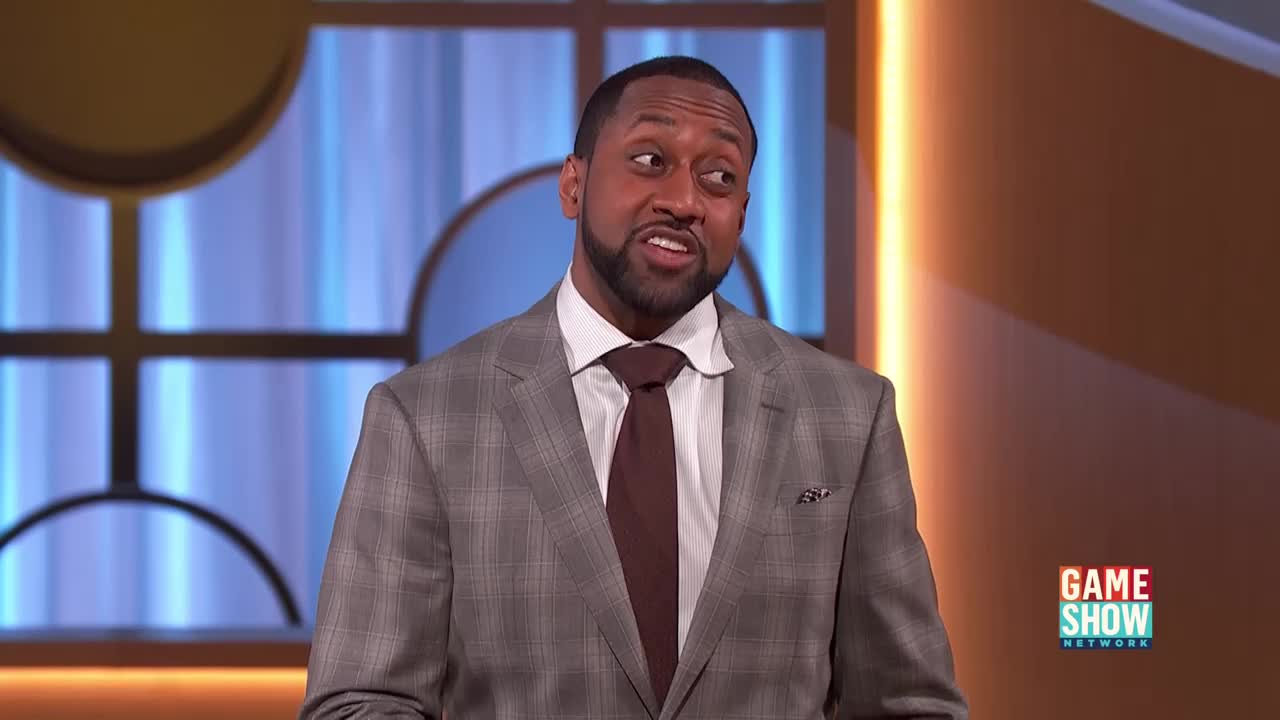 The man in the gray suit is speaking, his expression animated. A Game Show Network logo is visible in the background.
