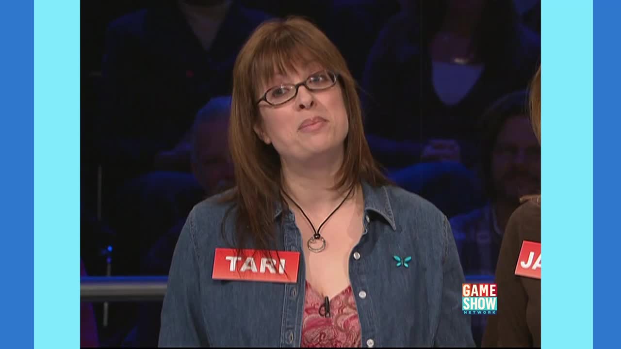 Tari, wearing a denim shirt and a name tag, is looking up with a slight smile on her face. The Game Show Network logo is visible on the right side of the frame.
