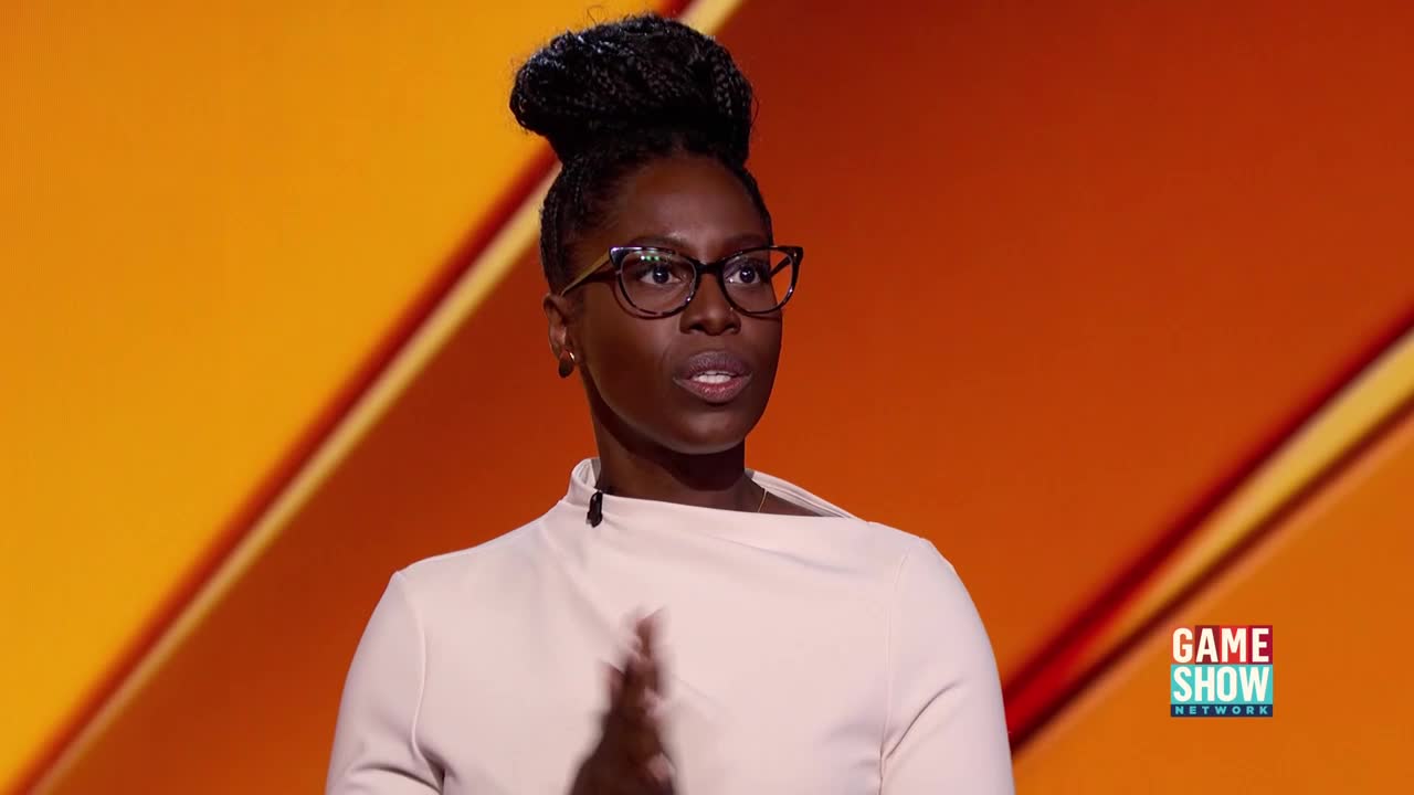 A woman in glasses and a light-colored top gestures with her hands as she speaks. Behind her, a Game Show Network logo sits against a bright orange backdrop.
