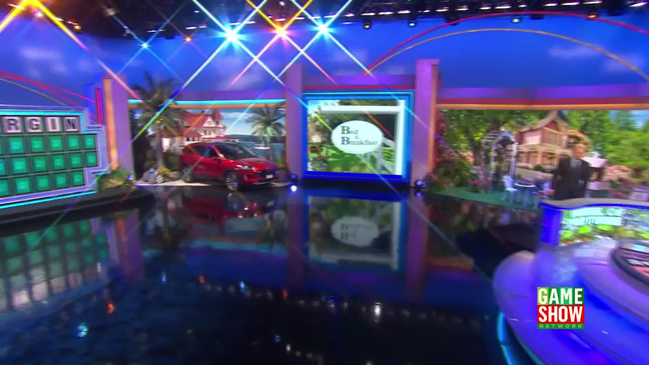 A red SUV sits on display next to a tropical backdrop, while a "Bed & Breakfast" graphic shines on a screen. The set of a game show, likely on Game Show Network, is brightly lit with a shiny, reflective floor.
