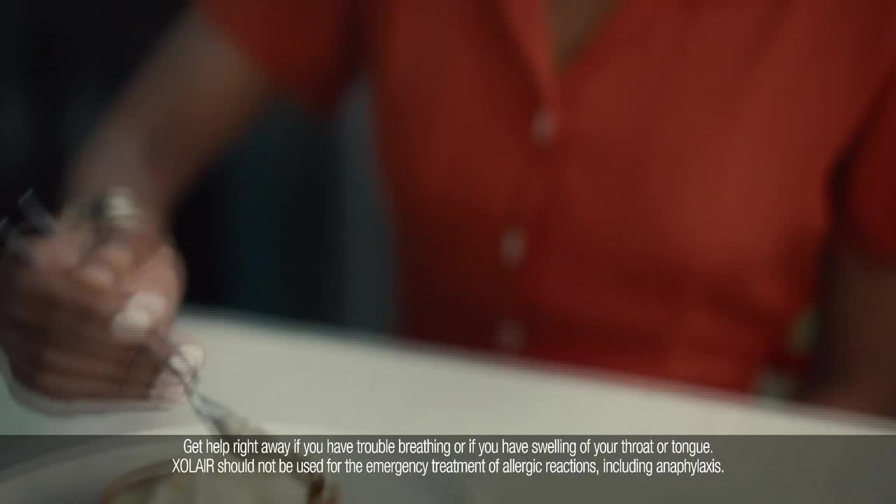 A hand, adorned with rings, holds a fork and moves it over a plate. The person wears a bright red shirt, and the scene is slightly out of focus.
