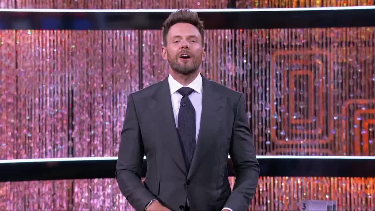 Joel McHale, in a dark suit and tie, stands center stage with his mouth open, likely about to speak. Behind him, a shimmering, beaded curtain provides the backdrop for a Game Show Network set.
