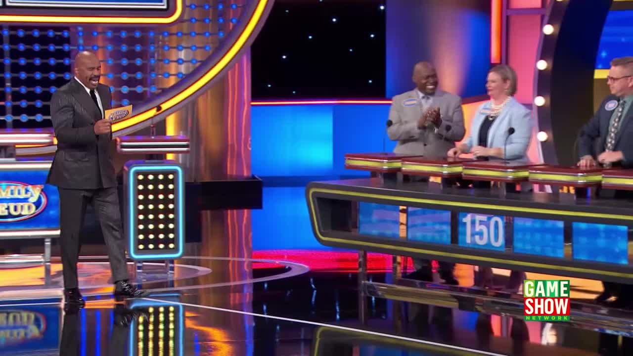 Steve Harvey, holding a card, stands on the "Family Feud" stage. The contestants, seated behind their podiums, watch, while one man claps.
