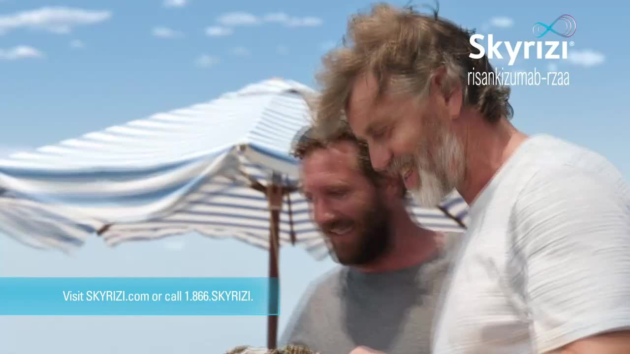 Two men, smiling, stand close together under a striped beach umbrella. They appear to be looking at something in their hands, with the ocean and sky in the background.
