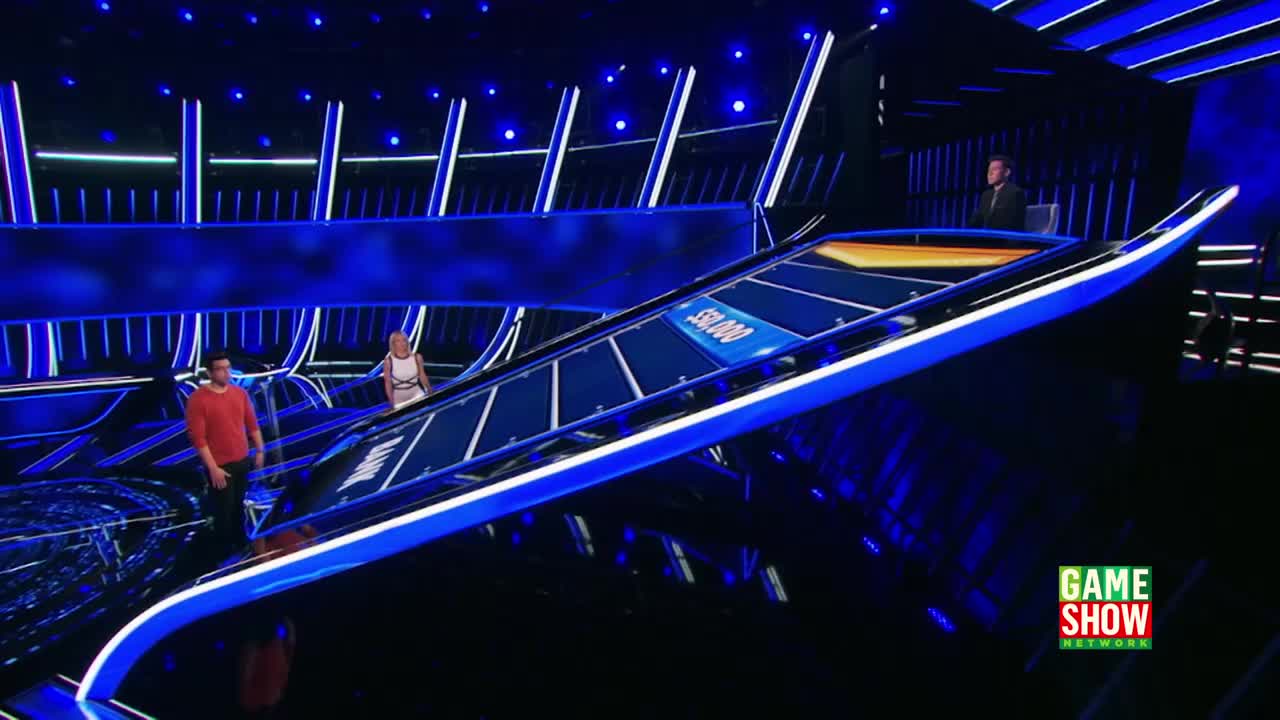 The contestants stand near a curved, illuminated platform, the set of a game show on the Game Show Network. A host in a suit observes the scene from a raised platform in the background.

