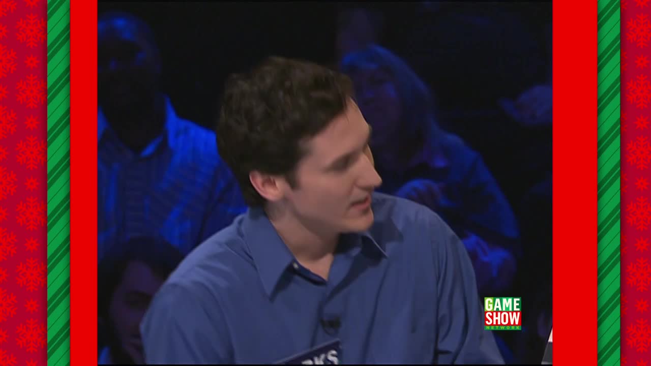 A man in a blue shirt is speaking, his gaze directed off-screen. The Game Show Network logo is visible in the corner, and the set has a festive, holiday-themed border.
