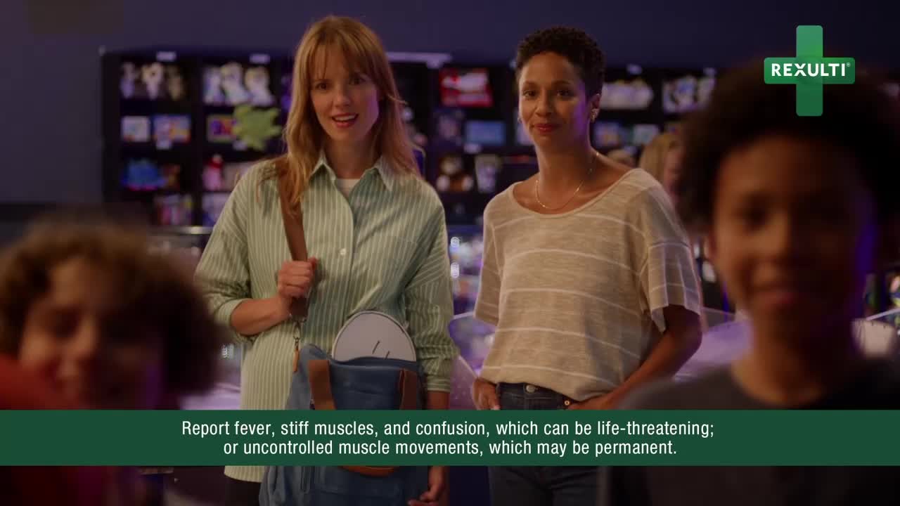 Two women stand and smile, likely in a public space, with a child's face in the foreground. The background is a bit blurry, but there's a vending machine visible.
