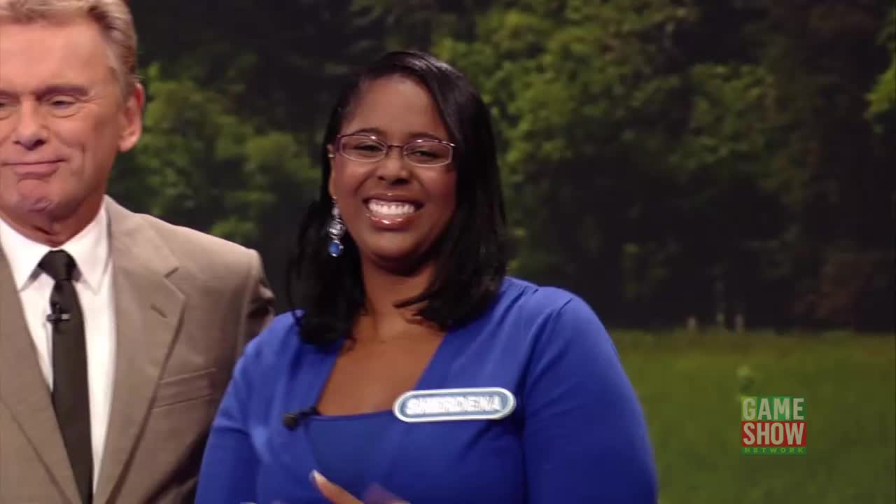The woman in the blue dress beams, her name tag reading "AMERIDENA." The host in the suit stands beside her, smiling, as they appear to be on a game show set.
